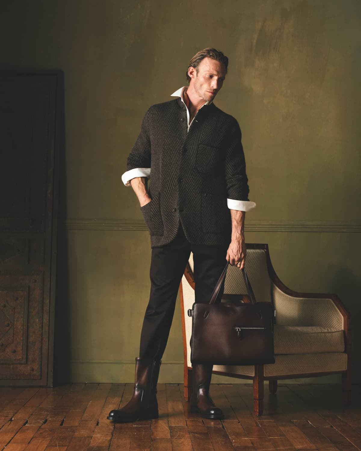 Model wearing dark Berluti tailoring and boots while holding a brown 1 Jour de Plus leather bag.