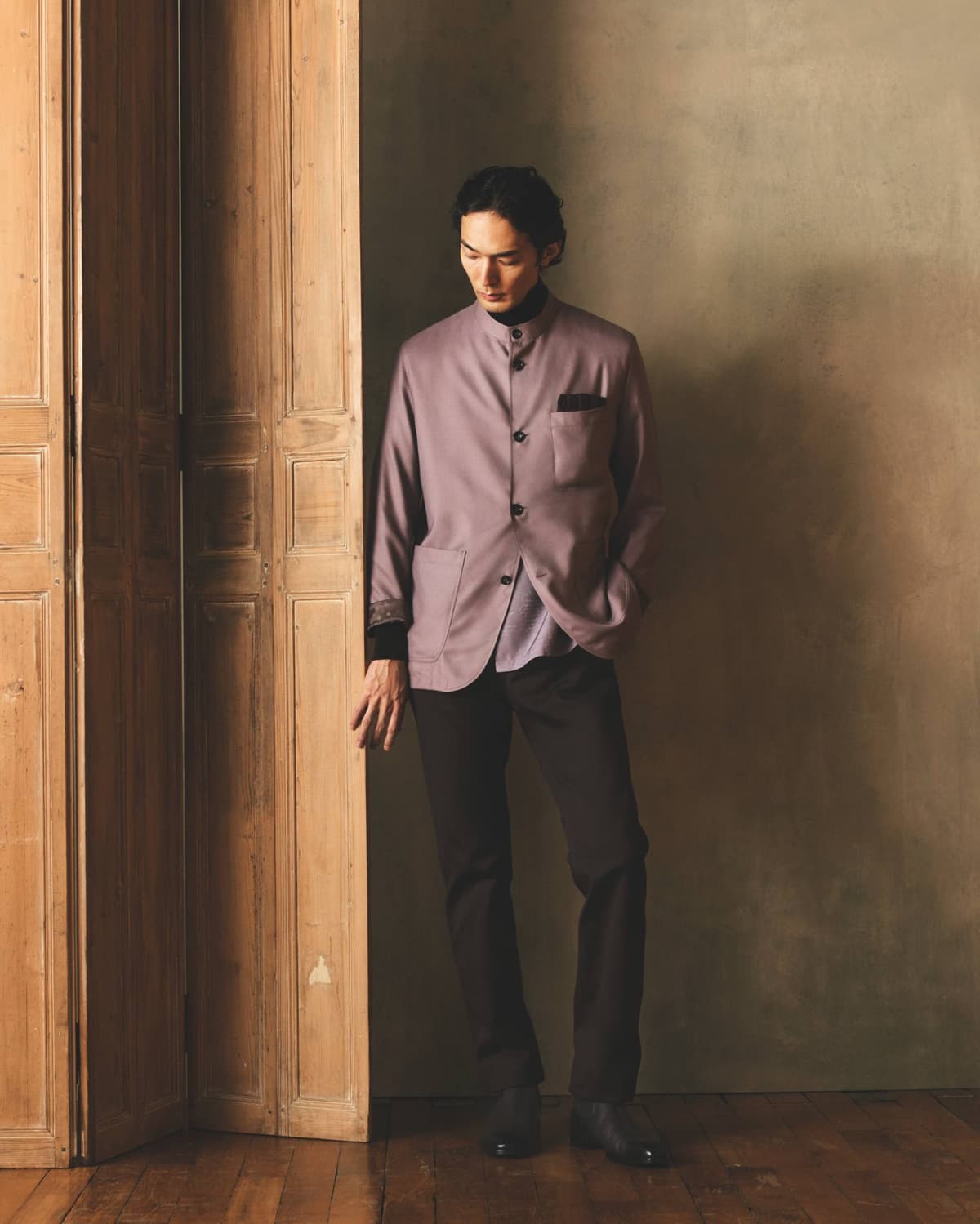 Model wearing a vintage pink Berluti Forestière jacket with dark trousers, part of Berluti Fall/Winter 2026 collection, beside a wooden doorway.
