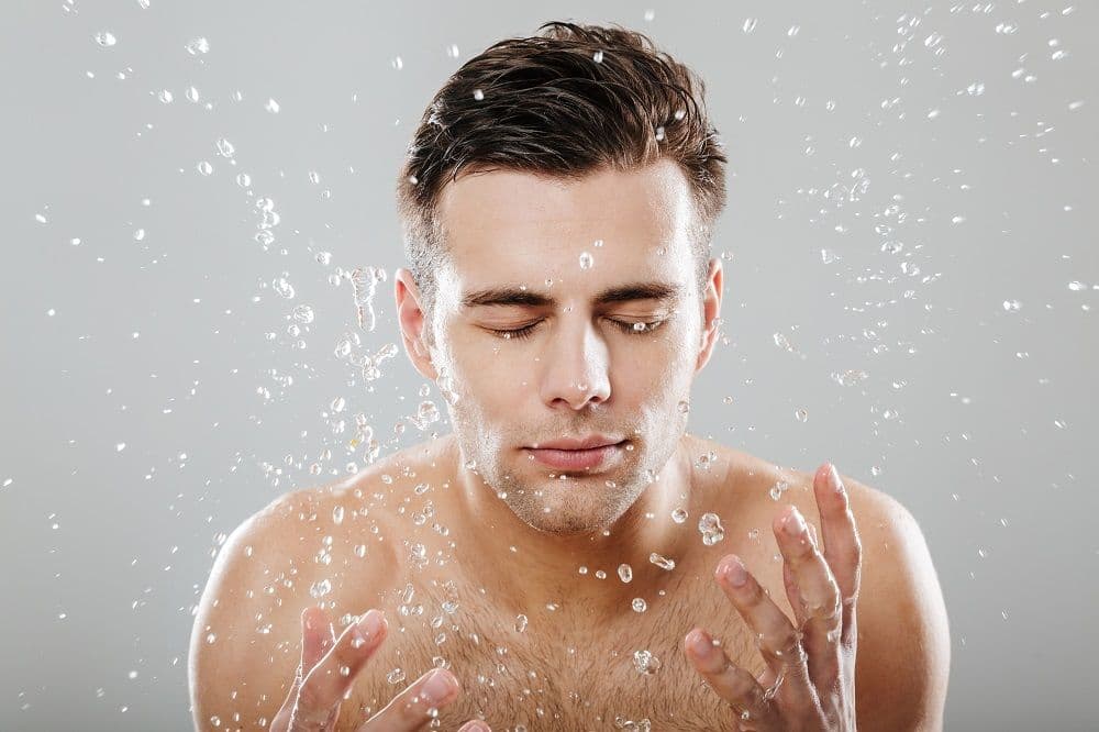 Close up portrait of a young half naked man surrounded by water drops washing his face isolated over gray background